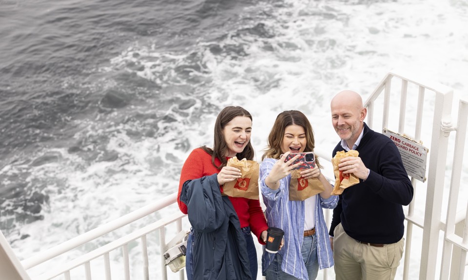 two ladies and a man taking a selfie out on deck with the sea behind them