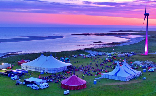 Festival tents and crowds on a grassy coastline at sunset, with a wind turbine and colorful sky in the background.