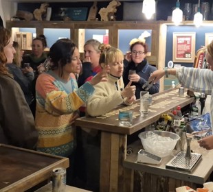 A busy bar scene with patrons gathered at the counter interacting with a bartender in a striped sweater, surrounded by wooden decor, hanging glasses.