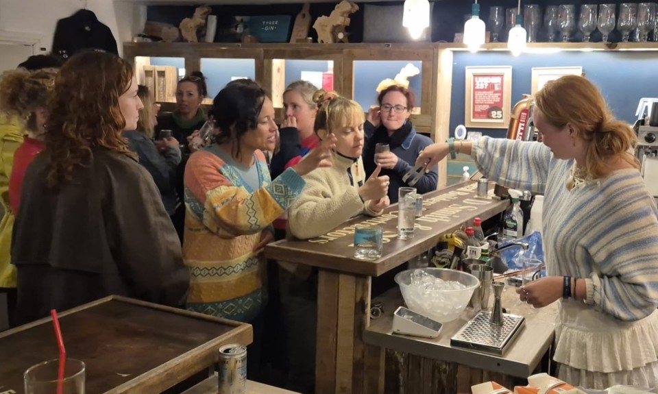 A busy bar scene with patrons gathered at the counter interacting with a bartender in a striped sweater, surrounded by wooden decor, hanging glasses.