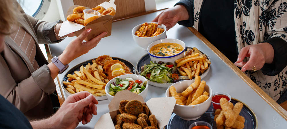 Three people dining on a ferry with a table full of food which includes fish and chips, salad, corn on the cob, onion rings and mozzarella sticks
