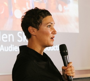 Speaker presenting at the Sea Change Film Festival, holding a microphone in front of a cinema audience presentation screen.
