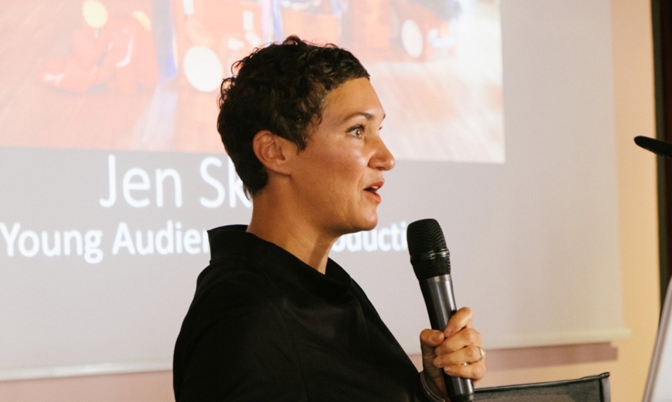 Speaker presenting at the Sea Change Film Festival, holding a microphone in front of a cinema audience presentation screen.