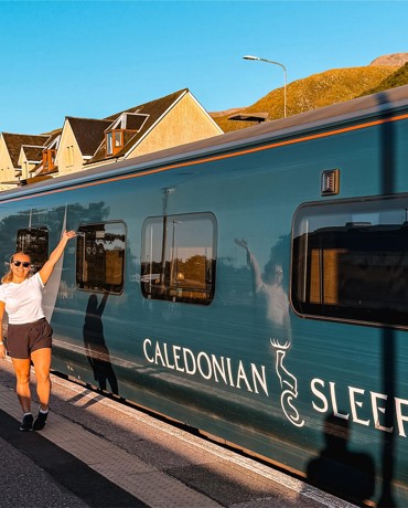Person in shorts and white shirt standing on train platform, reaching toward window of teal Caledonian Sleeper train.