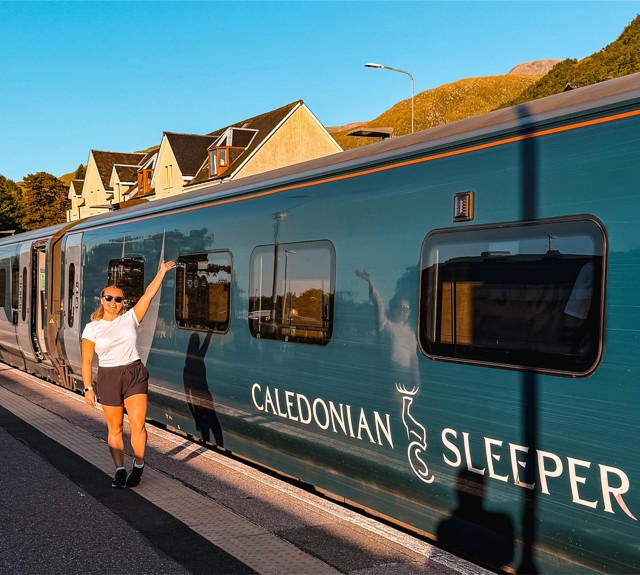 Person in shorts and white shirt standing on train platform, reaching toward window of teal Caledonian Sleeper train.