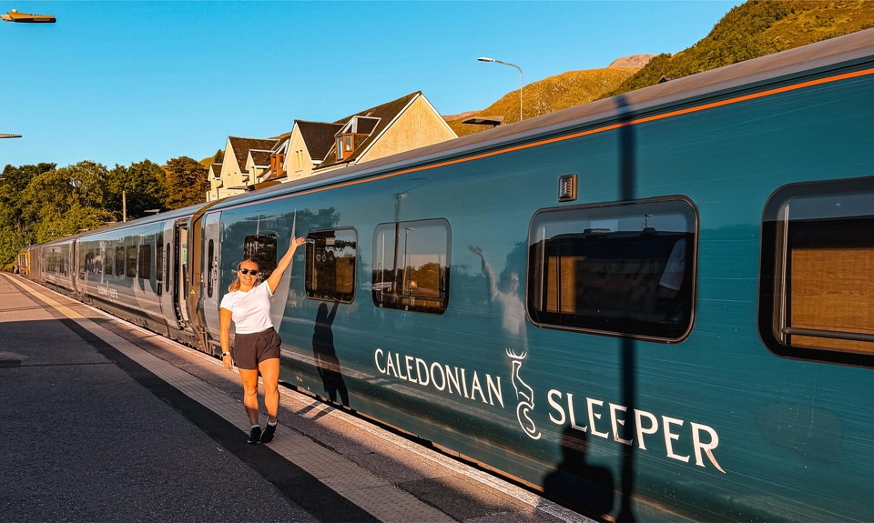 Person in shorts and white shirt standing on train platform, reaching toward window of teal Caledonian Sleeper train.