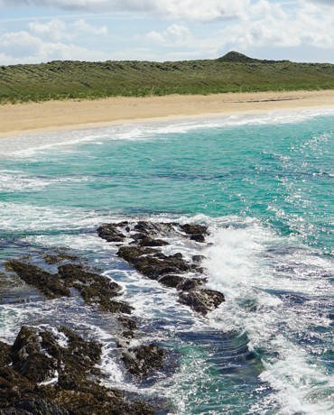 Turquoise waves break over dark rocks beside a long sandy beach backed by grassy dunes.