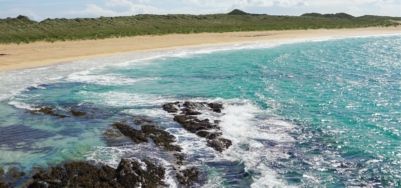 Turquoise waves break over dark rocks beside a long sandy beach backed by grassy dunes.