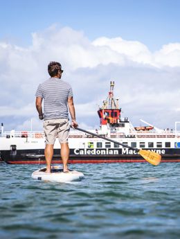 A man on the water on a stand up paddle board, admiring the CalMac ferry to Gigha