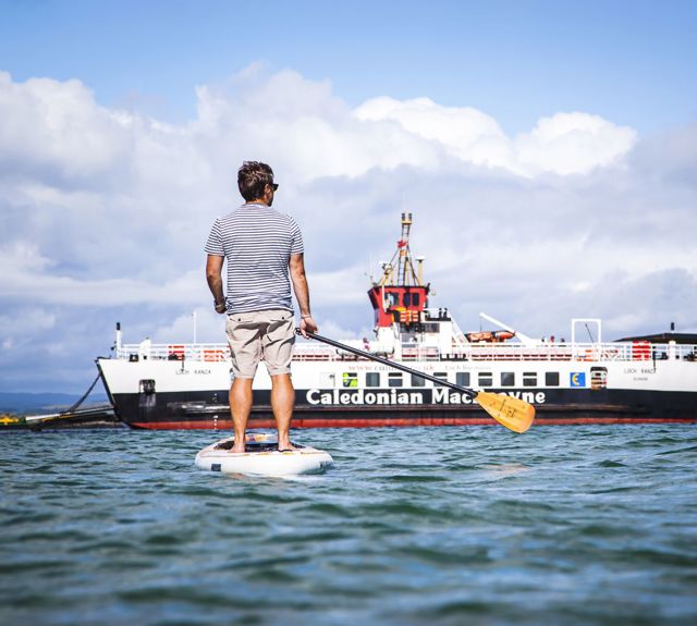 A man on the water on a stand up paddle board, admiring the CalMac ferry to Gigha