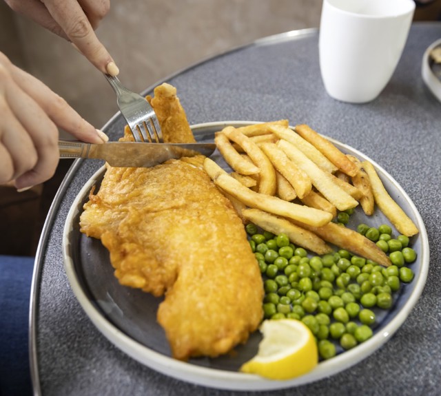 A plate with a large piece of battered fish topped with a lemon wedge, fries, and peas. The plate is on a blue table with utensils placed beside it.