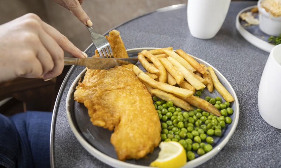 A plate with a large piece of battered fish topped with a lemon wedge, fries, and peas. The plate is on a blue table with utensils placed beside it.