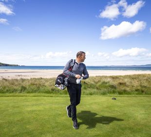 Golfer walking on a green course near the beach, with ocean waves and a clear blue sky in the background.