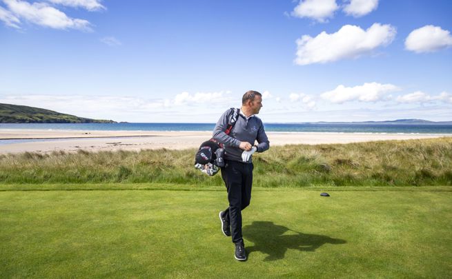 Golfer walking on a green course near the beach, with ocean waves and a clear blue sky in the background.