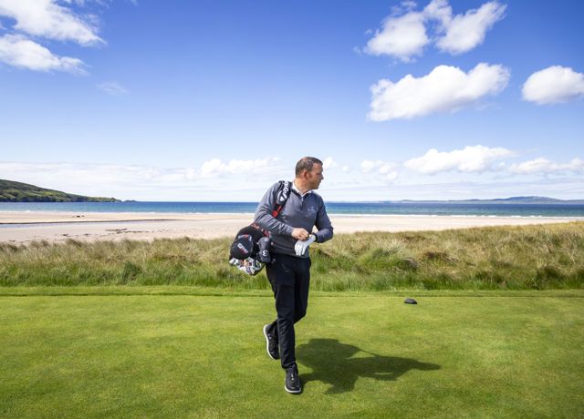 Golfer walking on a green course near the beach, with ocean waves and a clear blue sky in the background.