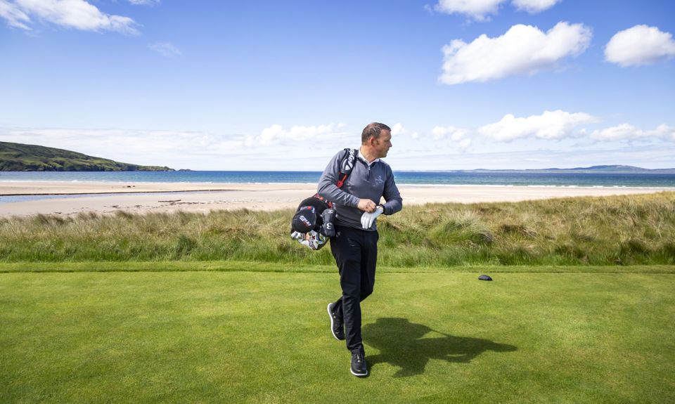 Golfer walking on a green course near the beach, with ocean waves and a clear blue sky in the background.