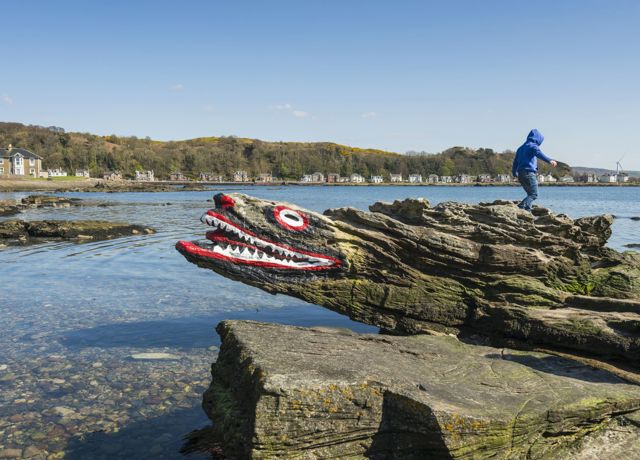 Child in blue jacket stands on "Crocodile Rock" - Rock formation at Millport seafront by the water, with houses and trees in the background.