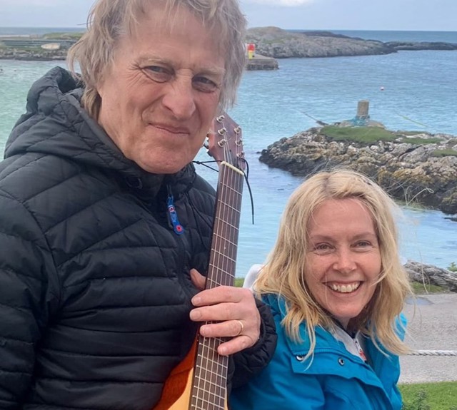 Two people by the sea, one holding a guitar, with rocky shore and boats in the background.