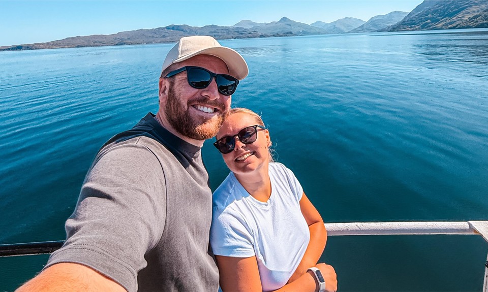 Two people on a boat with calm blue water and distant mountains in the background.