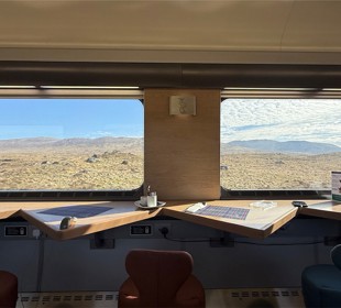 Train interior with tables, cups, and chairs facing large windows showing a desert landscape.