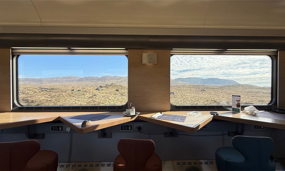 Train interior with tables, cups, and chairs facing large windows showing a desert landscape.