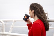 A female with long brown hair, wearing a red sweater, is standing on the deck holding a black cup with the CalMac logo. 