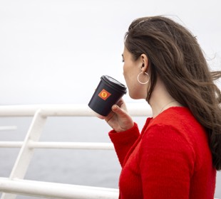 A female with long brown hair, wearing a red sweater, is standing on the deck holding a black cup with the CalMac logo. 