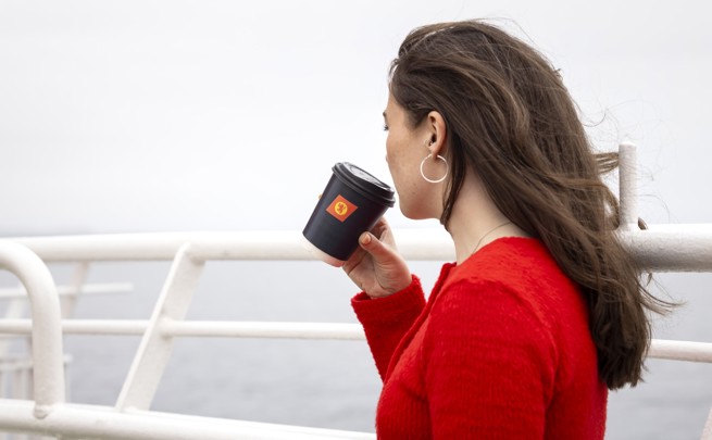 A female with long brown hair, wearing a red sweater, is standing on the deck holding a black cup with the CalMac logo. 
