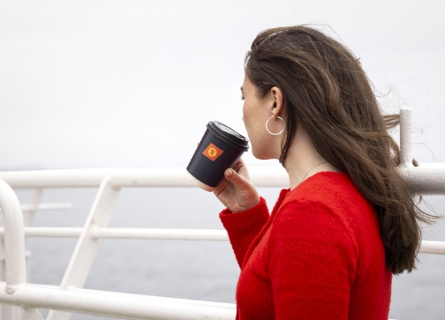 A female with long brown hair, wearing a red sweater, is standing on the deck holding a black cup with the CalMac logo. 