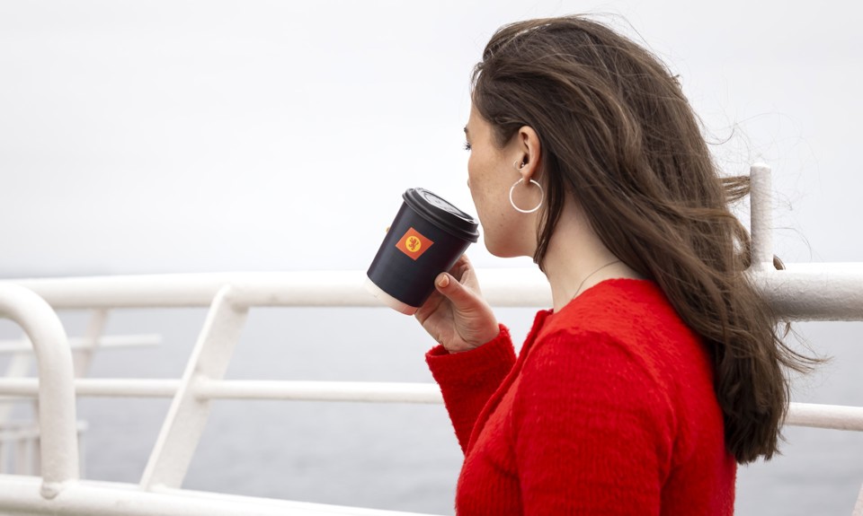 A female with long brown hair, wearing a red sweater, is standing on the deck holding a black cup with the CalMac logo. 