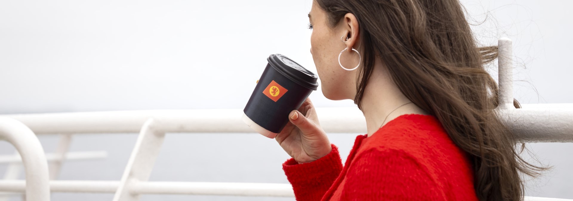 A female with long brown hair, wearing a red sweater, is standing on the deck holding a black cup with the CalMac logo. 