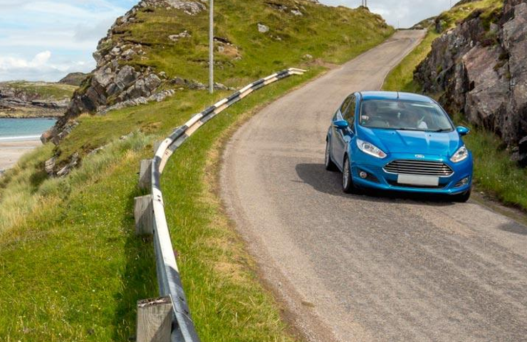 A blue car driving down a single track, coastal road