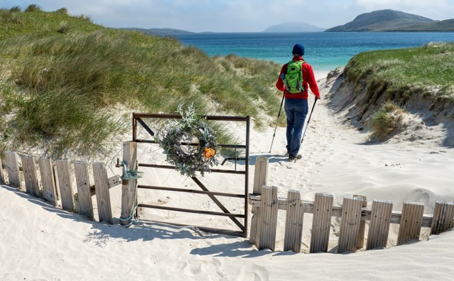 Person with a green backpack walking through sandy dunes toward a turquoise sea, passing a gate decorated with a wreath.