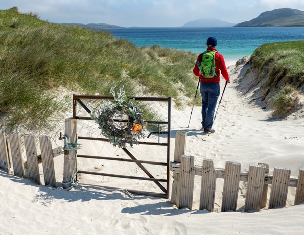 Person with a green backpack walking through sandy dunes toward a turquoise sea, passing a gate decorated with a wreath.