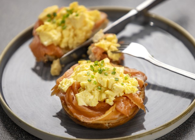 someone cutting into a bagel topped with scrambled egg and smoked salmon with a knife and fork