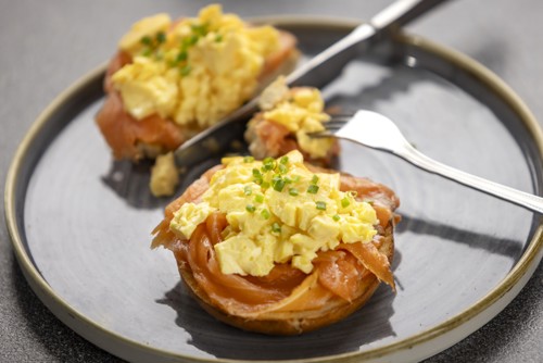 Someone cutting a bagel with smoked salmon and scrambled egg with a knife and fork