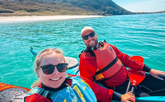 Two people in life jackets kayak on clear turquoise water near a sandy beach with mountains in the background.
