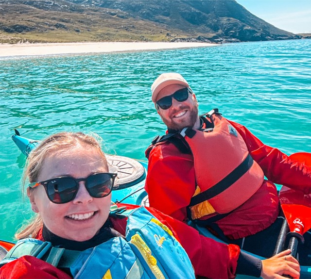 Two people in life jackets kayak on clear turquoise water near a sandy beach with mountains in the background.
