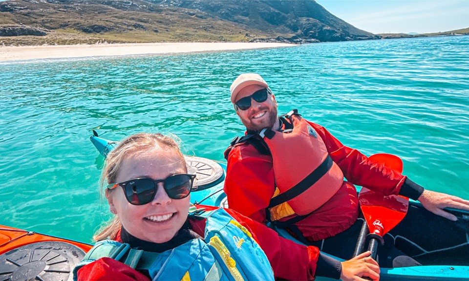 Two people in life jackets kayak on clear turquoise water near a sandy beach with mountains in the background.