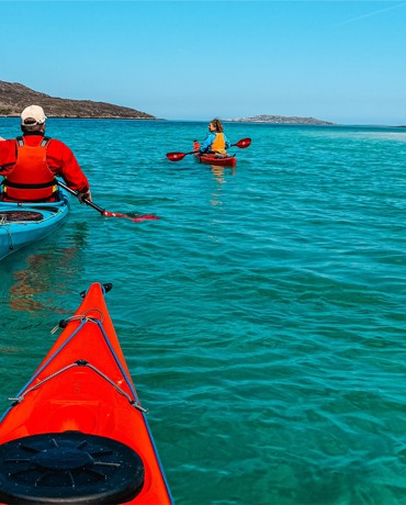 Two people kayaking in clear turquoise water near sandy beach with rocky hills in background.