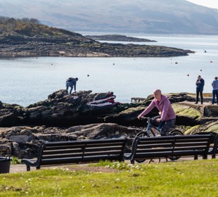 people playing on top of a crocodile shaped rock on the beach with a cyclist riding past