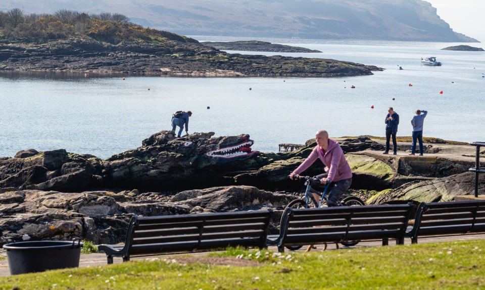 people playing on top of a crocodile shaped rock on the beach with a cyclist riding past