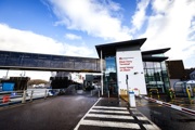 Oban Ferry Terminal entrance with English and Gaelic signage, a skybridge, and a ferry docked under a partly cloudy sky.