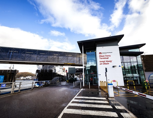 Oban Ferry Terminal entrance with English and Gaelic signage, a skybridge, and a ferry docked under a partly cloudy sky.