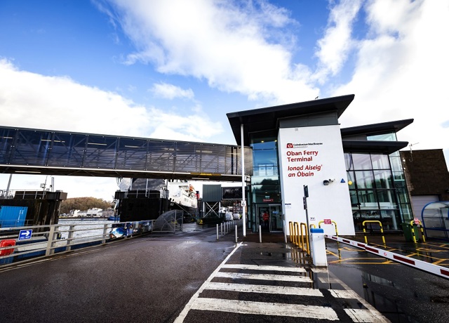 Oban Ferry Terminal entrance with English and Gaelic signage, a skybridge, and a ferry docked under a partly cloudy sky.