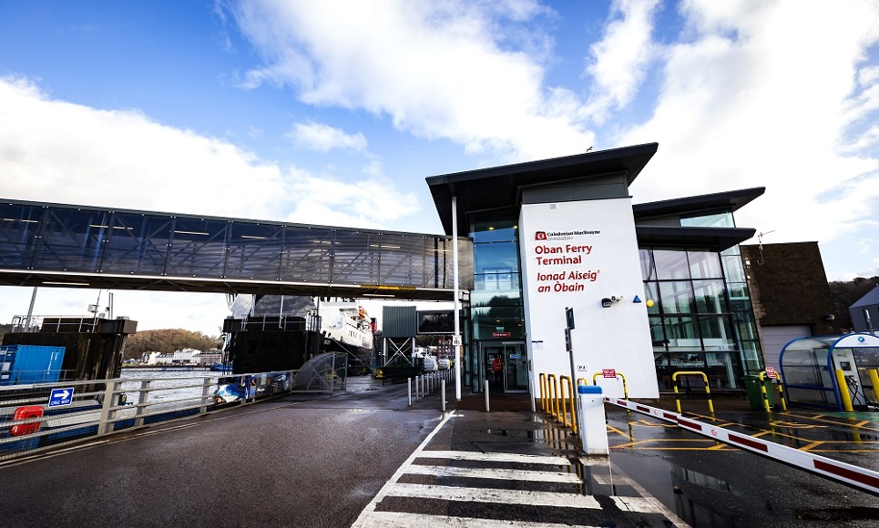 Oban Ferry Terminal entrance with English and Gaelic signage, a skybridge, and a ferry docked under a partly cloudy sky.