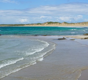 White sandy beach with gentle waves, turquoise water and dune-backed shoreline beneath a bright sky.