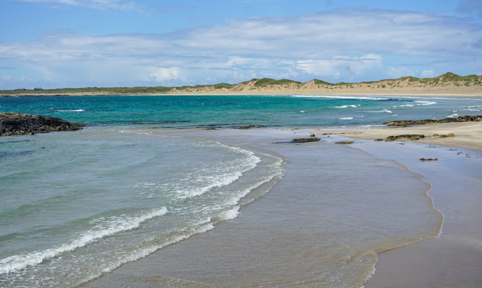 White sandy beach with gentle waves, turquoise water and dune-backed shoreline beneath a bright sky.