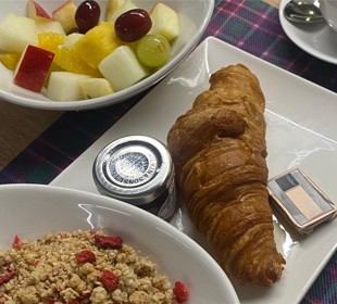Breakfast with croissant, jam, butter, fruit salad, granola with berries, and coffee on a plaid placemat