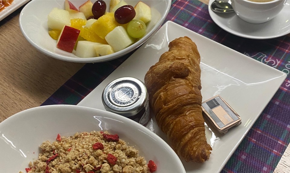 Breakfast with croissant, jam, butter, fruit salad, granola with berries, and coffee on a plaid placemat